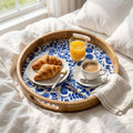 A round wooden serving tray with a blue floral patterned liner sits on a bed with white linens. The tray holds croissants on a small plate, a glass of orange juice, a cup of coffee on a saucer, and a folded napkin.