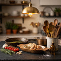 A warm, rustic kitchen scene with a brass pendant light glowing above a stone countertop. A cast-iron skillet with asparagus, sliced bread on a wooden board, garlic, cherry tomatoes, rosemary, wooden utensils in a ceramic container, and a small amber oil bottle are arranged on the counter.