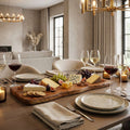 A beautifully styled dining table set for a dinner gathering, featuring a large wooden charcuterie and cheese board with assorted cheeses, crackers, grapes, and figs, alongside wine glasses and plates in a warm, modern living room setting.